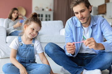 Obraz premium Happy father smilling daughter lying on warm floor enjoying creative activity, drawing pencils coloring pictures in albums, mother resting on couch, family spend free time together.