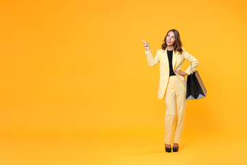 Full length portrait of smiling young woman in suit jacket hold package bags with purchases after shopping point finger aside up on mock up copy space isolated on yellow background. Black friday sale.