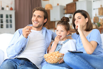 Happy family with child sitting on sofa watching tv and eating popcorn, young parents embracing daughter relaxing on couch together.