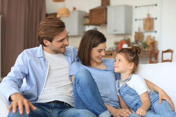 Positive friendly young parents with smiling little daughter sitting on sofa together while relaxing at home on weekend.
