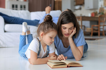 Mother and little daughter read a book together, lying on the floor in the living room.