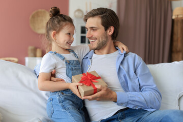 Cute kid daughter make surprise to daddy, little girl presents gift box to father sit on sofa....