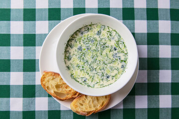 Popular spring or summer cold soup okroshka. Okroshka in a white bowl, bread and herbs on a table.