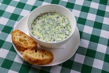 Popular spring or summer cold soup okroshka. Okroshka in a white bowl, bread and herbs on a table.