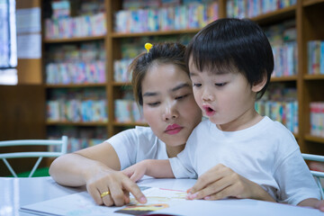 Asian young mother with little boy reading book together in library. Happy family. Parent educating children. Pre-school learning concept.
