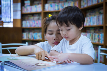 Asian young mother with little boy reading book together in library. Happy family. Parent educating children. Pre-school learning concept.