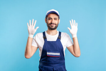 Naklejka premium Cheerful handyman in overalls raising hands with surgical gloves, smiling to camera. Safety clothes, protection and hygiene in service industry, courier delivery, housekeeping maintenance. studio shot