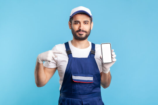 Workman In Overalls And Gloves Pointing Mobile Phone With Mock Up Blank Display, Advertising Area For Online Delivery Order App, House Repair Maintenance Services. Indoor Studio Shot Isolated On Blue