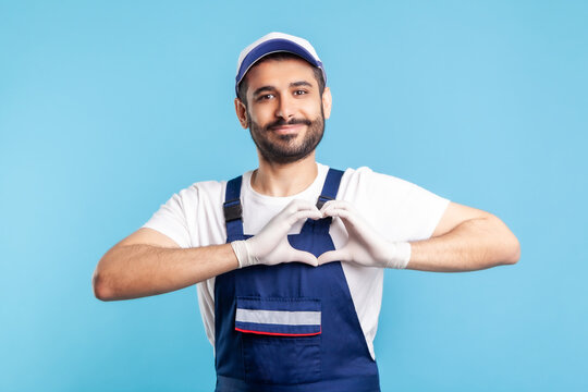 Love our customers! Handyman in overalls and hygiene gloves showing heart gesture and smiling friendly to camera. Profession of service industry, courier delivery, housekeeping maintenance. isolated