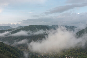 Morning fog over a mountain valley in autumn. Miniature houses. Green and golden trees. Sky with clouds.