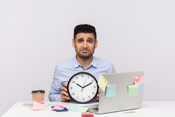 Overtime work. Unhappy exhausted man employee sitting in office workplace, holding clock and frowning upset, sticky notes all around reminding of deadline. studio shot isolated on white background