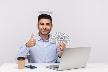 Excellent online earnings! Angelic rich businessman with nimbus on head holding dollars and showing thumbs up, like gesture, sitting at laptop workplace. studio shot isolated on white background