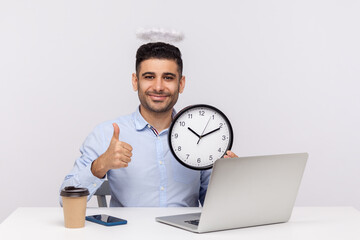 Time management. Joyful punctual man employee with angelic nimbus sitting in office workplace, holding clock and showing thumbs up, happy to meet deadline. studio shot isolated on white background