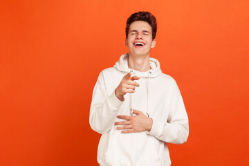 Happy pleased young man in white hoodie pointing finger at you and sincerely laughing holding hand...