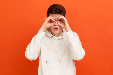 Funny curious young man in casual white sweatshirt looking far away through binoculars gesture, spying with open mouth, shocking content. Indoor studio shot isolated on orange background