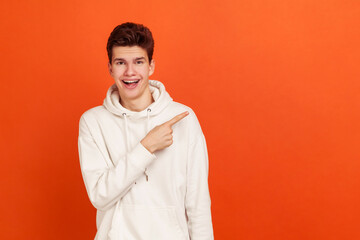 Extremely cheerful young man in casual white hoodie pointing finger to right demonstrating empty...