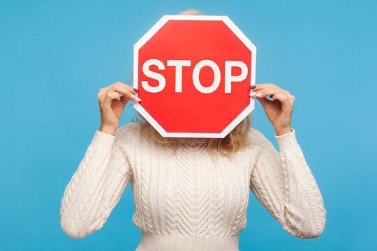 Woman in white knitted sweater hiding her face behind stop sign, afraid talking about domestic violence, scared victim. Indoor studio shot isolated on blue background