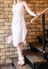 A beautiful slender girl is standing on a wooden staircase in a gentle pink dress. Model with heels in leather sandals. holds a stylish bag in the color of the image. Woman on brick wall background