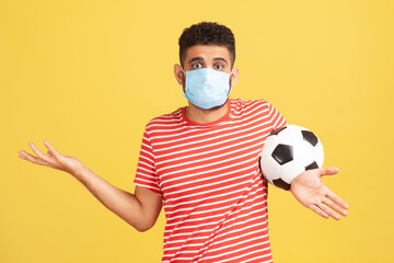 Frustrated uncertain man in red t-shirt and hygienic face mask spreading hands holding ball, confused how to play football safety. Indoor studio shot isolated on yellow background