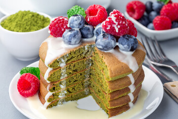 Cuted stack of matcha pancakes served with condensed milk, blueberry and raspberry on a white plate, horizontal