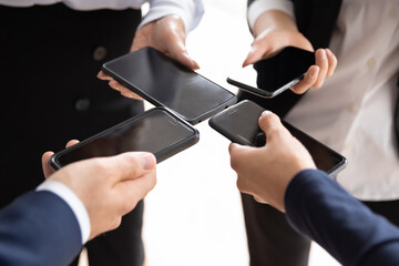 Crop close up of diverse businesspeople hold smartphones communicate via application in office. Colleague coworkers use modern cellphone gadgets at meeting at workplace. Technology, mobile concept.