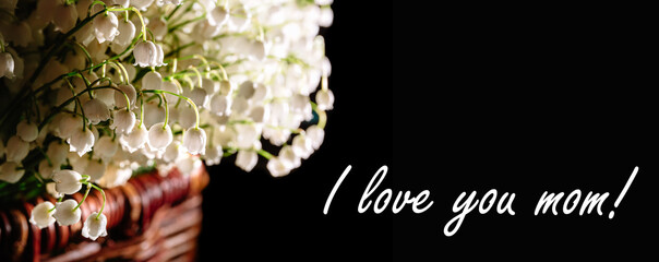 Postcard with the phrase I love you mom. Banner with large beautiful bouquet of white lilies of the valley, packed in a brown wicker basket and isolated on a black background