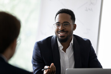 Smiling African American young businessman in glasses talk with business partner at office meeting....
