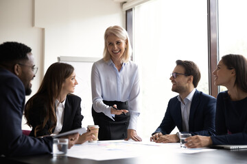 Smiling middle-aged businesswoman hold lead meeting with diverse colleagues discuss ideas together. Happy multiracial businesspeople have fun brainstorming at team office briefing. Teamwork concept.
