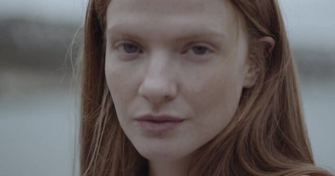 Close up Portrait of beautiful serene redhead woman looking at camera on the beach, ginger fashion model female on beach headshot with moody sky.