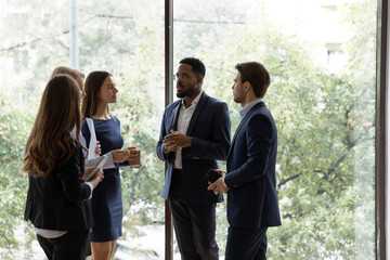 Multiracial colleagues stand in office hallway drink coffee talk brainstorm at work break together....