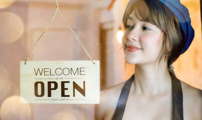 Business owner attractive young Asian woman in apron hanging we're open sign on front door smiling...