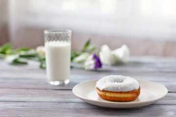 White donut on a plate with a cup of coffee on a wooden table. Light flowers lie nearby