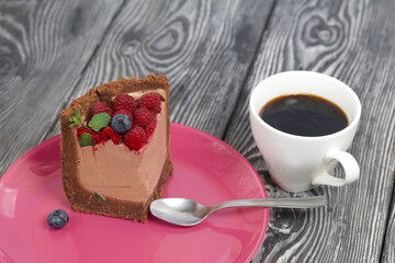 A piece of no-bake chocolate cake with cream cheese. Garnished with raspberries, blueberries and mint leaves. Nearby is a cup of coffee. On the surface of pine boards painted in black and white.