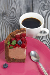 A piece of no-bake chocolate cake with cream cheese. Garnished with raspberries, blueberries and mint leaves. Nearby is a cup of coffee. On the surface of pine boards painted in black and white.