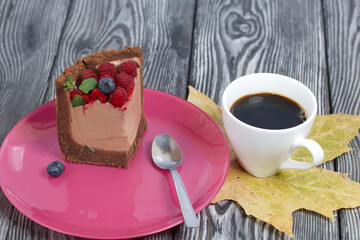 A piece of no-bake chocolate cake with cream cheese. Garnished with raspberries, blueberries and mint leaves. Nearby is a cup of coffee and dried maple leaves.
