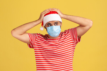 Fototapeta na wymiar Unhappy sad man in red t-shirt and santa claus hat with surgical medical mask on face touching head, migraine, fear of contracting coronavirus. Indoor studio shot isolated on yellow background