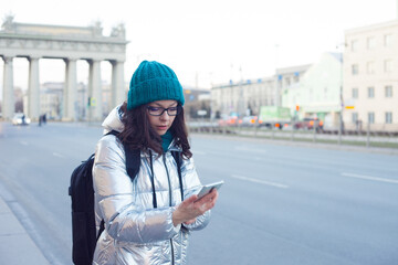 girl in a down jacket and hat stands at a public transport stop and uses a smartphone.