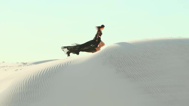 Silhouette of a happy woman running in the desert. Long black silk dress and hair flying fluttering in the wind in slow motion. A tourist is enjoying a vacation in the Dubai desert. Video footage 4K