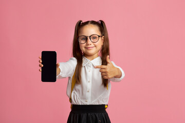 Gadgets for study. Schoolgirl in glasses, school uniform shows a finger at blank screen of smartphone