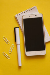 Creative flat lay, photo of workspace desk with smartphone, notepad, a pen, some paper clips, copy space, yellow background, minimal style, selective focus