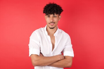 Coquettish Young arab man with afro hair wearing shirt standing on red wall, smiling happily, blinking at camera in a playful manner, flirting with you. Human facial expressions and emotions