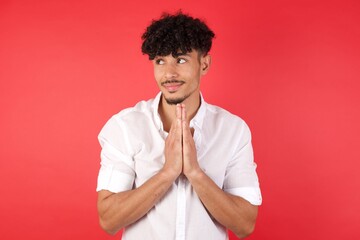 Young arab man with afro hair wearing shirt standing over isolated red background making up plan in mind holding hands together, setting up an idea.