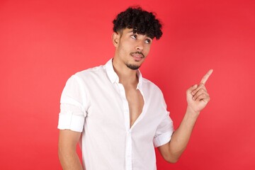 Smiling Young arab man with afro hair wearing shirt standing over isolated red background pointing up and looking at the camera