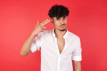 Unhappy  Young arab man with afro hair wearing shirt standing over isolated red background imitates gun shoot makes suicide gesture keeps two fingers on temples has puzzled expression 