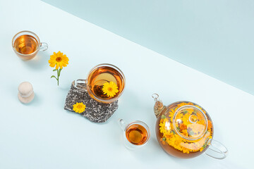 Cups of herbal tea, transparent teapot with calendula flowers and stones on blue background....
