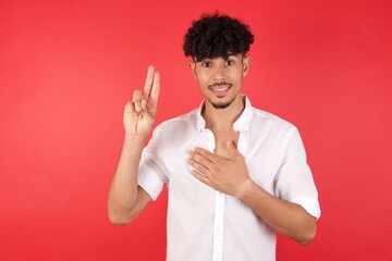 Young arab man with afro hair wearing shirt standing over isolated red background smiling swearing...