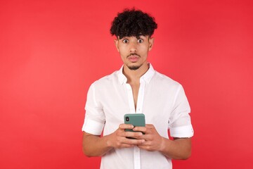 Young arab man with afro hair wearing shirt standing over isolated red background opens mouth hold telephone reading advert unbelievable big shopping prices .