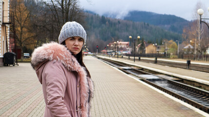 Fototapeta na wymiar A woman in a gray knitted hat and a pink jacket with fur is waiting for the train at the station looking at the arriving train. Rail transportation. Autumn and winter travel concept.