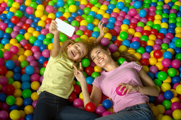 Fototapeta na wymiar Young mom with her daughter taking selfie while playing in ball pond at indoor amusement park, above view