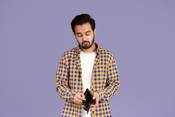 Economic crisis. Unhappy Indian guy looking into empty wallet on violet background